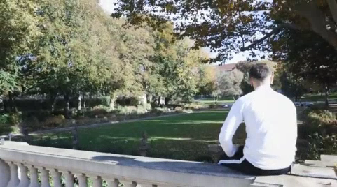 Thumbnail_Azubi_Samer / Azubi Samer sitzt auf einem Geländer. Im Hintergrund eine große Wiese.