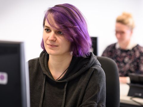 junge Frau mit lila Haaren sitzt vor einem Monitor