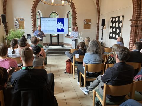 Zwei Menschen stehen auf Bühne und halten Vortrag, davor Menschen im Publikum