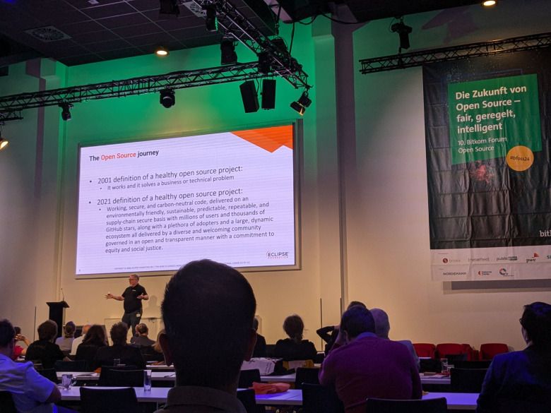 Publikum schaut auf eine Bühne auf der eine Person einen Vortrag hält