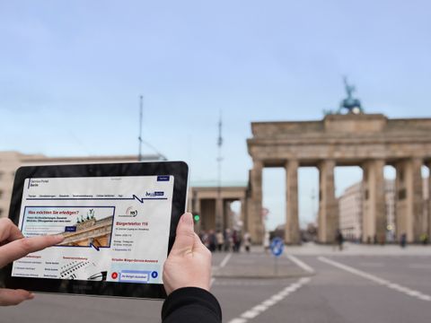 Das Brandenburger Tor im Hintergrund und im vordergrund und ein hochgehaltenes Tablet mit einer website