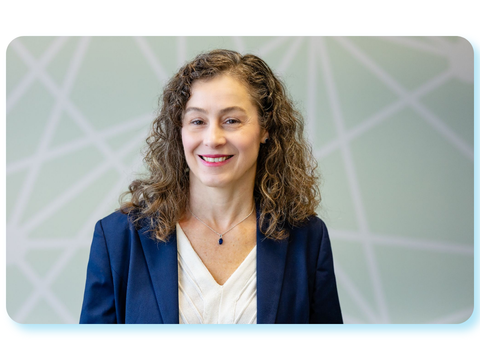 Porträtfoto von Dr. Maria Borelli. Sie lächelt in die Kamera und trägt einen dunkelblauen Blazer über einer hellen Bluse vor einem hellgrünen Hintergrund mit geometrischem Muster.