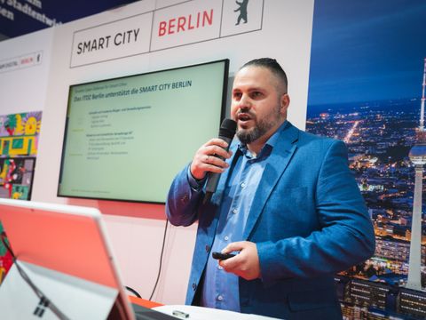 Marcel Bilal spricht auf einer Messe oder Veranstaltung in ein Mikrofon. Im Hintergrund sind weitere Personen sowie ein digitaler Bildschirm am Stand des ITDZ Berlin zu sehen.