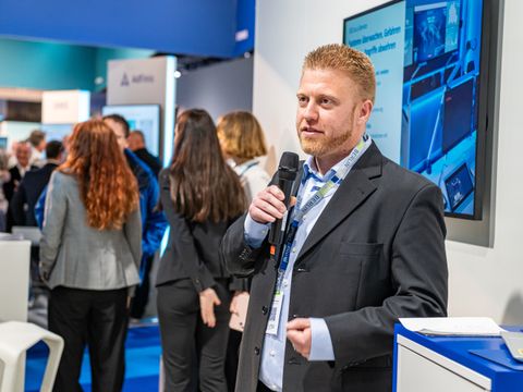 Maximilian Schneider spricht auf einer Messe oder Veranstaltung in ein Mikrofon. Im Hintergrund sind weitere Personen sowie ein digitaler Bildschirm am Stand des ITDZ Berlin zu sehen.