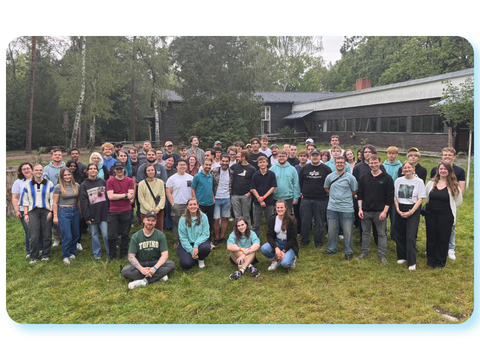 Gruppenfoto von Auszubildenden und Studierenden des ITDZ Berlin. Die Gruppe steht und sitzt draußen auf einer Wiese vor einem Gebäude, umgeben von Bäumen.
