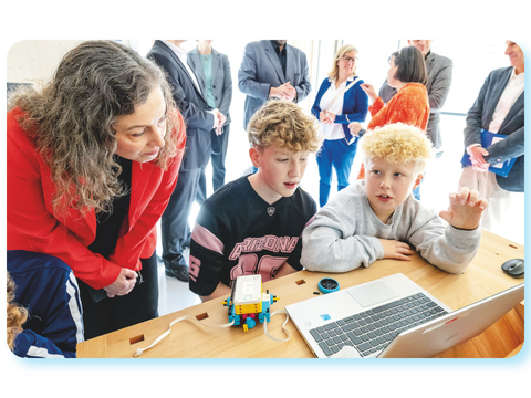 Kinder arbeiten gemeinsam an einem Laptop und einem kleinen Robotik-Modell, während eine Erwachsene zuschaut.