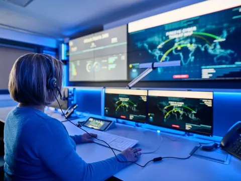 Eine Mitarbeiterin sitzt am Schreibtisch an Maus und Tastatur. Sie hat zwei kleine und zwei große Monitore weiter entfernt vor sich.