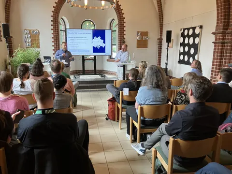 Zwei Menschen stehen auf Bühne und halten Vortrag, davor Menschen im Publikum