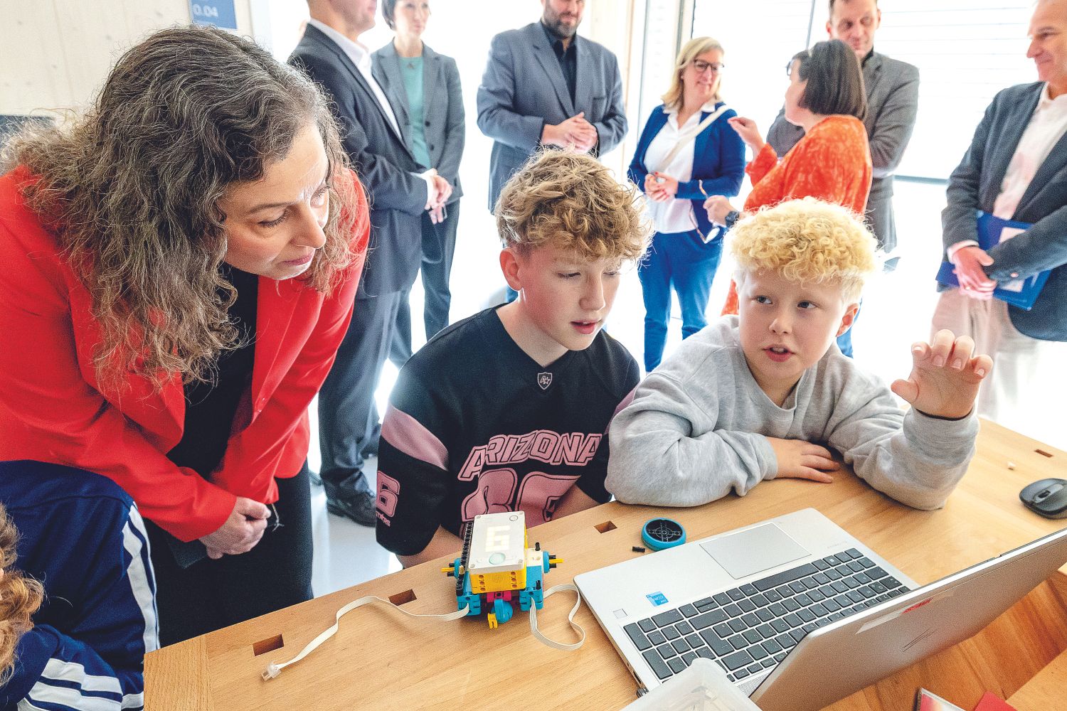 Kinder arbeiten gemeinsam an einem Laptop und einem kleinen Robotik-Modell, während eine Erwachsene zuschaut.