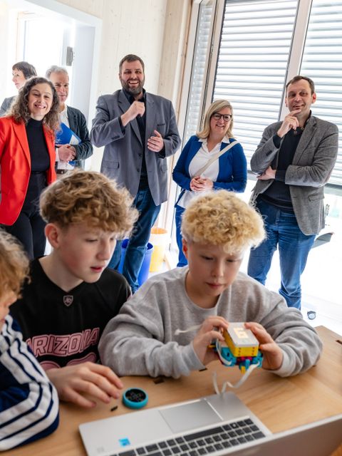 Im Vordergrund Schüler vor einem Laptop, im Hintergrund Erwachsene, die lächelnd zuschauen