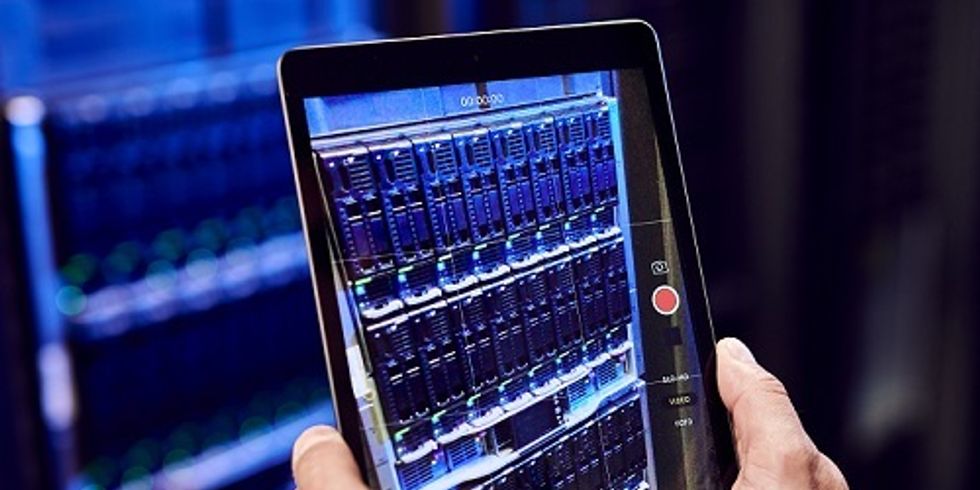 ein verschwommenes Serverboard im Hintergrund ist gestochen scharf auf einem Tablet abgebildet, welches von zwei Händen gehalten wird