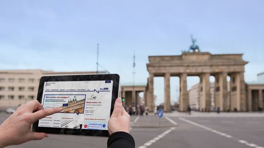 Das Brandenburger Tor im Hintergrund und im vordergrund und ein hochgehaltenes Tablet mit einer website