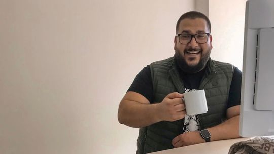 Serhat, Mitarbeitender des ITDZ Berlin steht an seinem Schreibtisch und hält lavhend eine Tasse Kaffee in der Hand