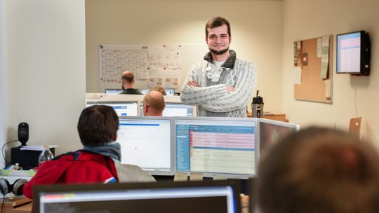 Ein Mitarbeiter steht in einem Büro und lächelt ins Bild mit mehreren MItarbeitern die an Computern arbeiten.