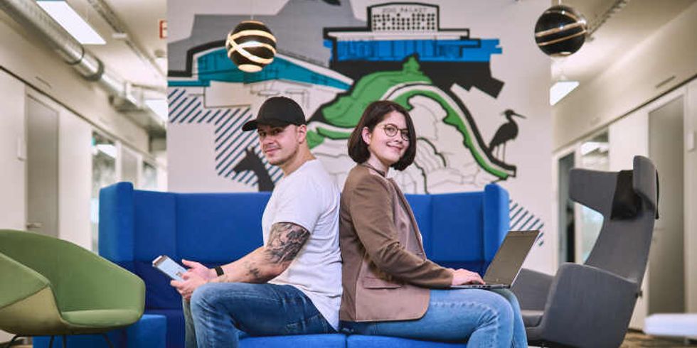 Zwei Leute mit einem Computer und einem Handy in der Hand gucken in die Kamera.